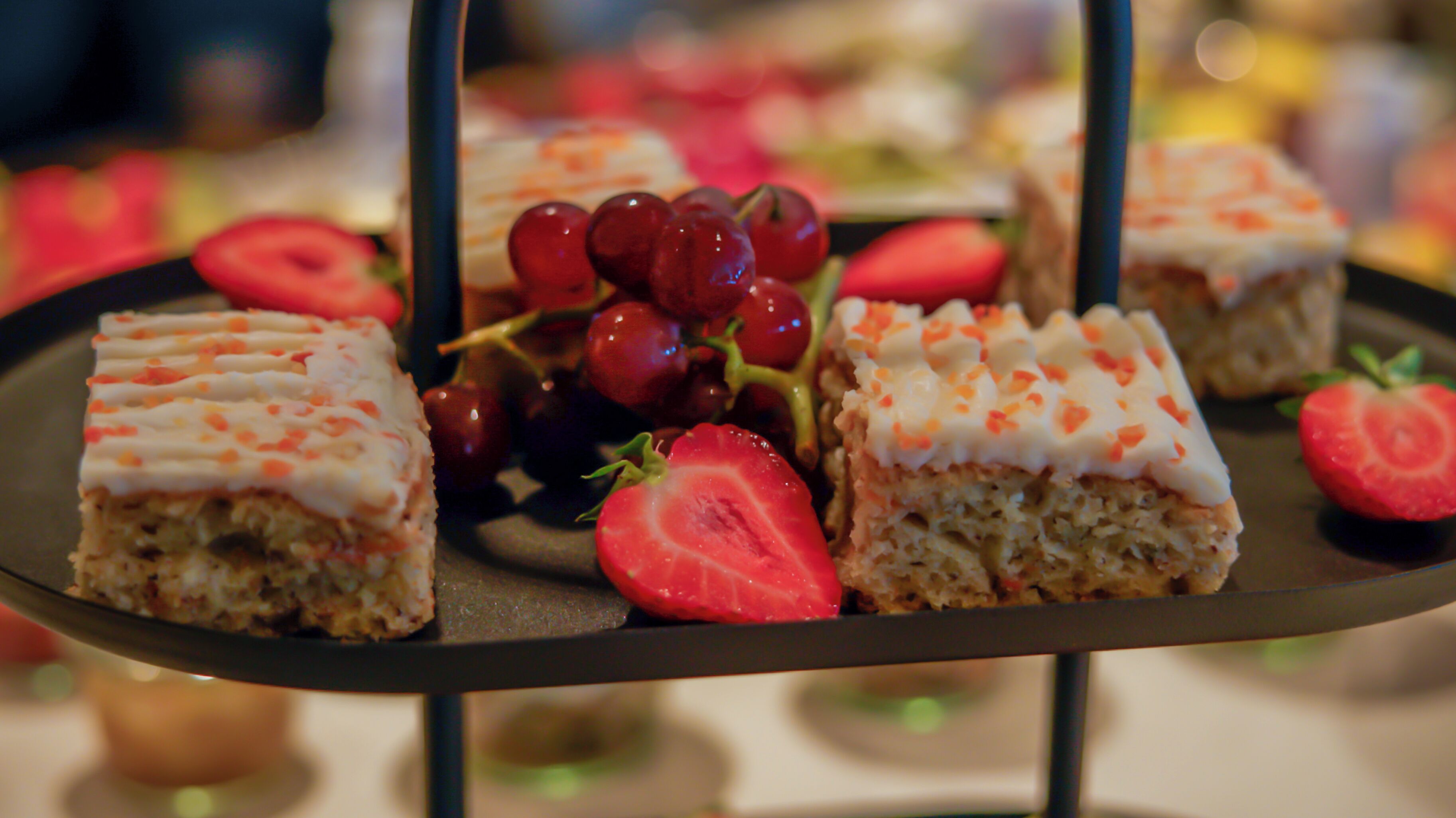 Eine Dessertplatte mit quadratischen Kuchen, belegt mit weißem Zuckerguss und orangenen Streuseln, dazu frische Erdbeeren und rote Trauben.