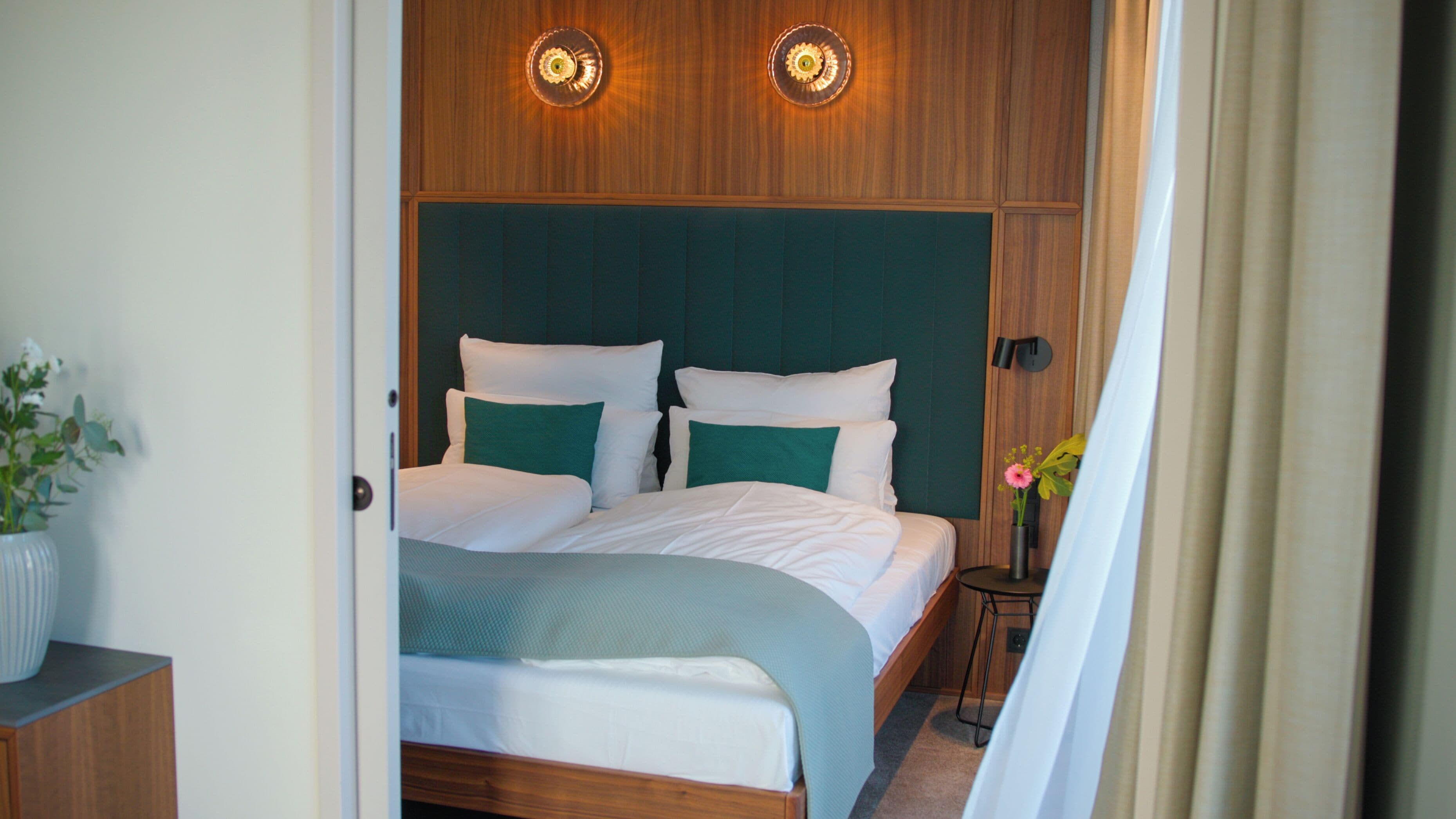 A neatly made bed with white and green bedding, a wooden headboard, and two wall-mounted lights.