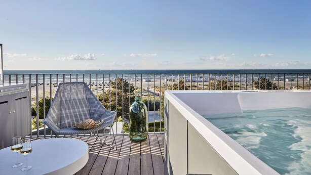 Un balcón con vista a la playa, una bañera de hidromasaje y una zona de estar con una mesa de vidrio y dos vasos.