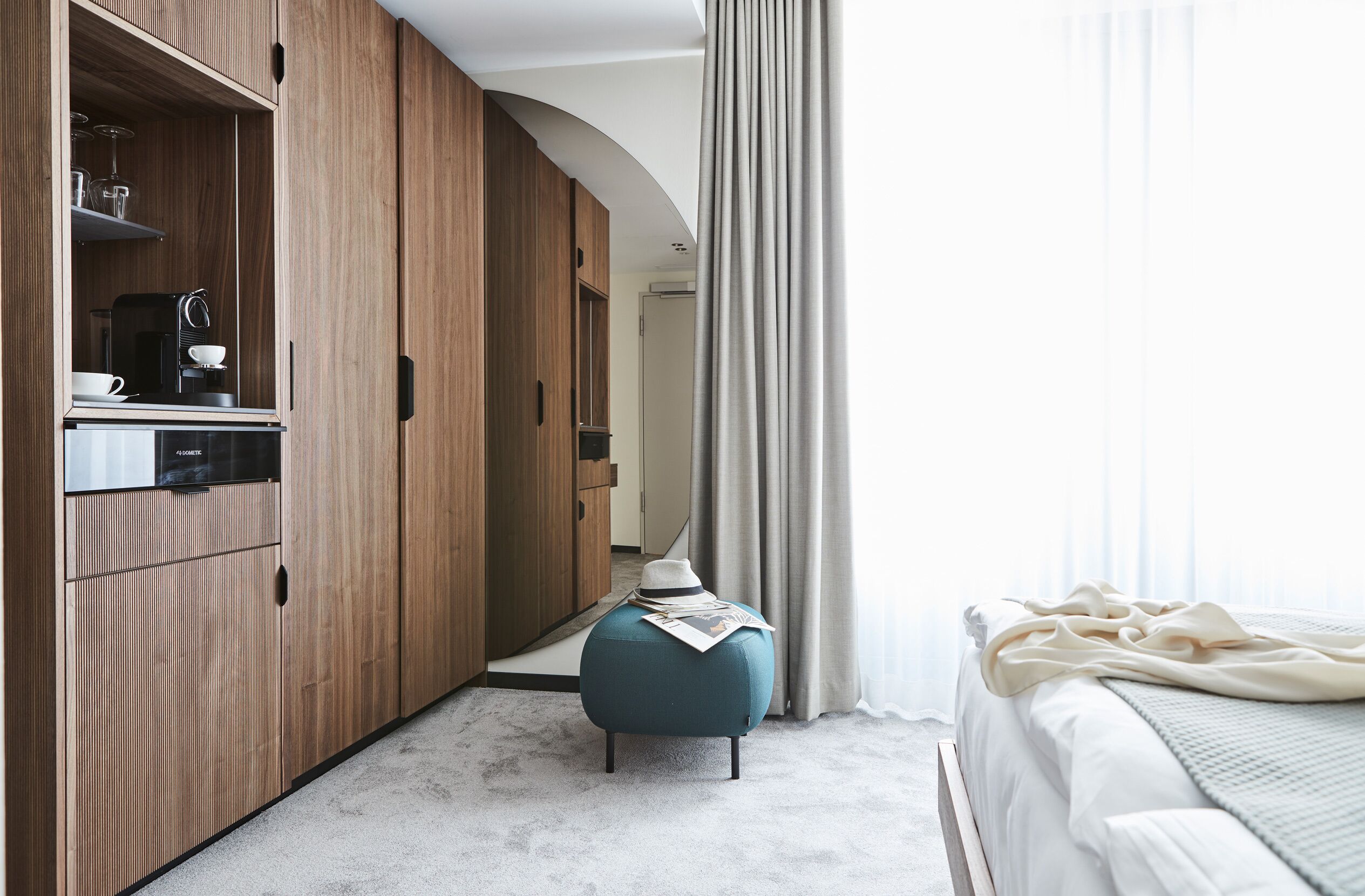 A bedroom with a bed, a wooden wardrobe, a kitchenette, and a window with curtains.
