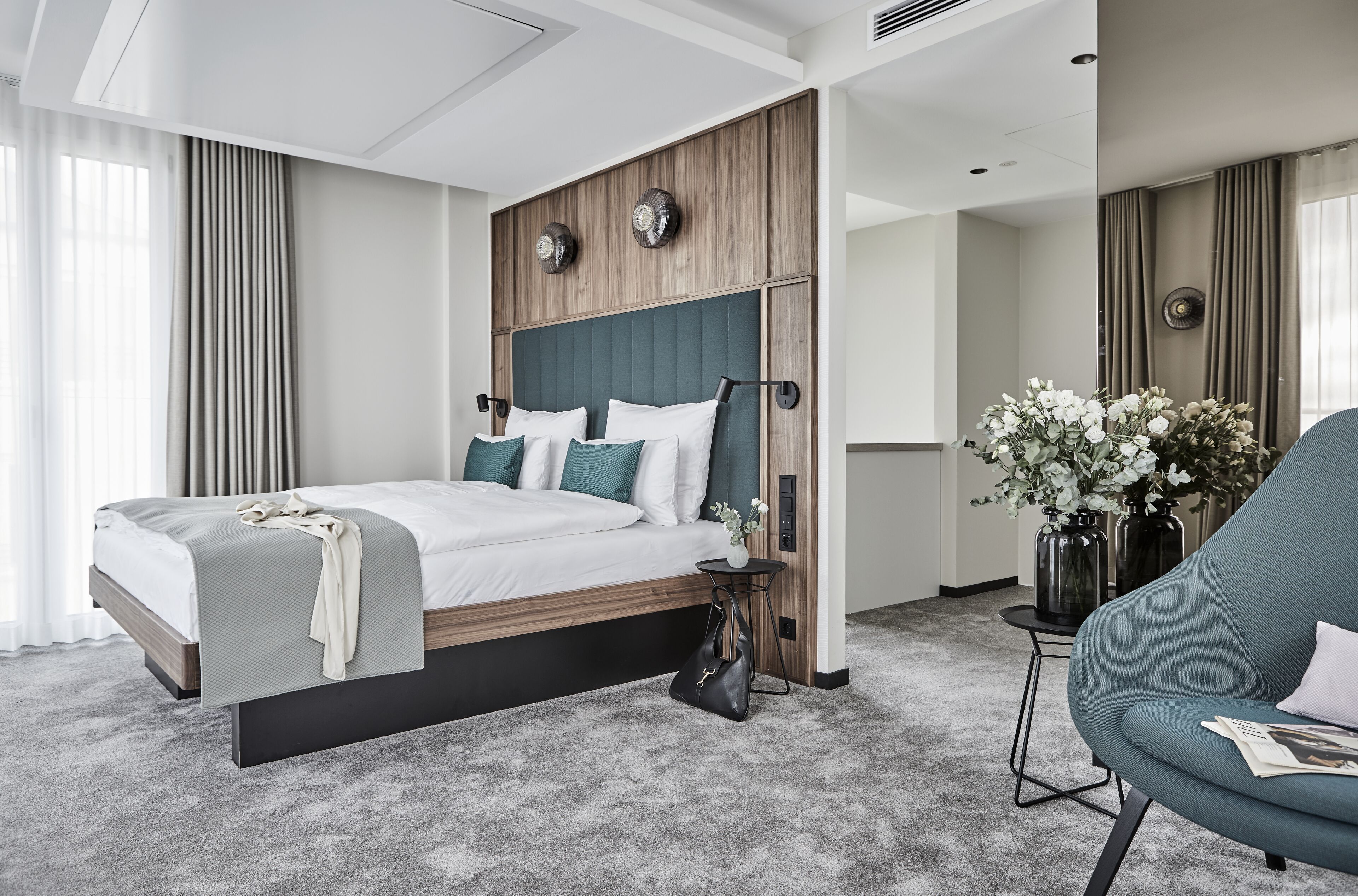 A modern hotel room with a large bed, a side table, a chair, and a vase with flowers.