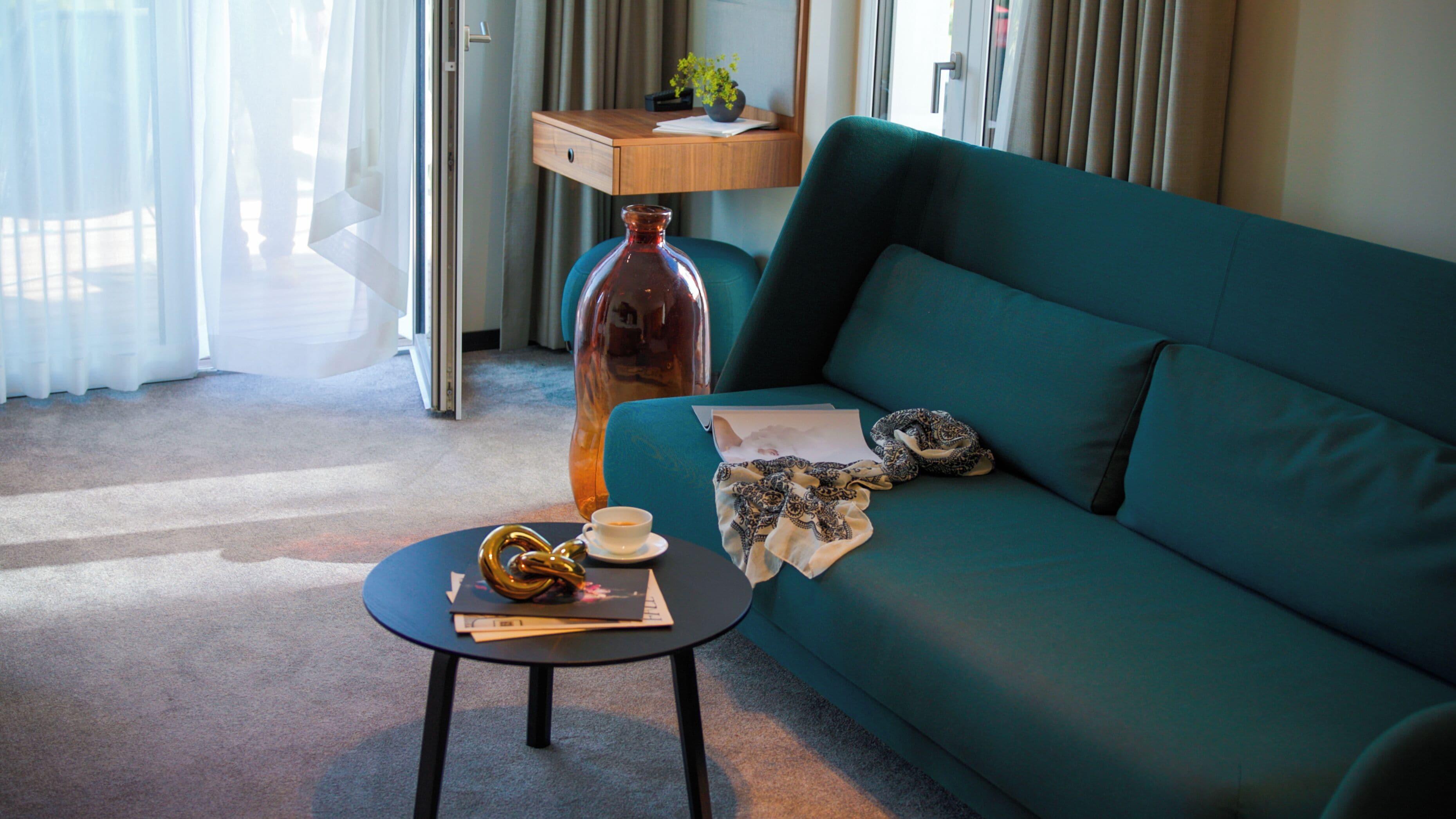 A teal sofa, a small round table with a book and a cup, a decorative vase, and a lamp on a wooden shelf.