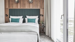 Un dormitorio con cabecera de madera, cama con ropa de cama blanca y azul turquesa, una mesita con un florero y vistas al exterior a través de una puerta corrediza.