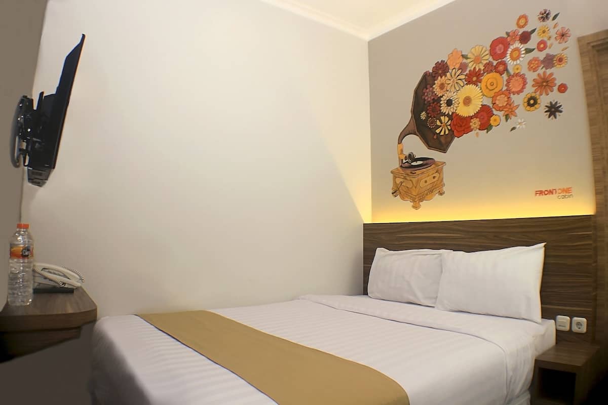 A hotel room with a bed, a bedside table with a bottle and a phone, a wall-mounted TV, and a decorative wall art piece.