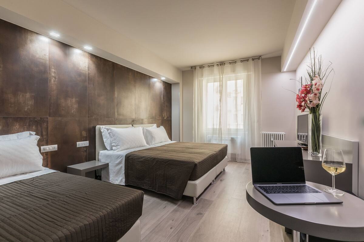 Una habitación de hotel moderna con dos camas, una laptop sobre una mesita y un florero con flores.