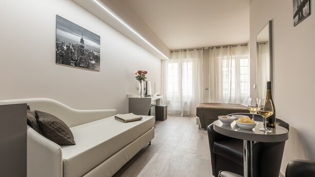 Een moderne hotelkamer met een bank, een eettafel met een fles wijn en glazen, een klein bijzettafeltje en een stadsgezichtschilderij aan de muur.