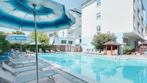 A swimming pool with lounge chairs and umbrellas, surrounded by a building with balconies. - Hotel Foglieri (Cesenatico)