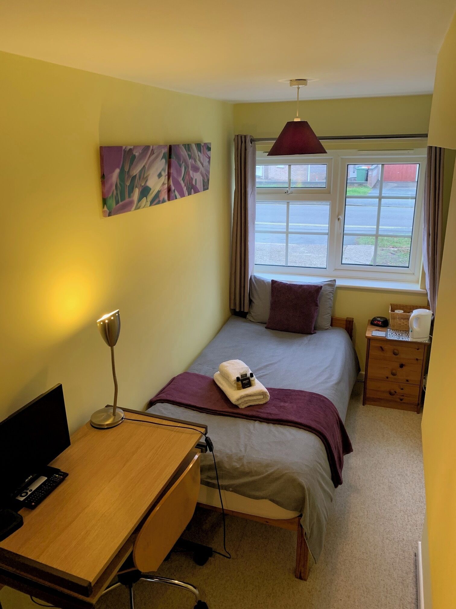 Habitación de hotel con cama, escritorio, silla, lámpara, mesita de noche, ventana y un cuadro en la pared.
