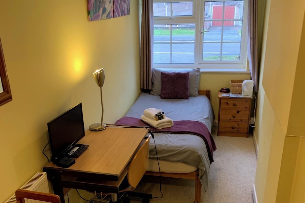 Una habitación pequeña con una cama, mesita de noche, lámpara y un escritorio con una computadora.