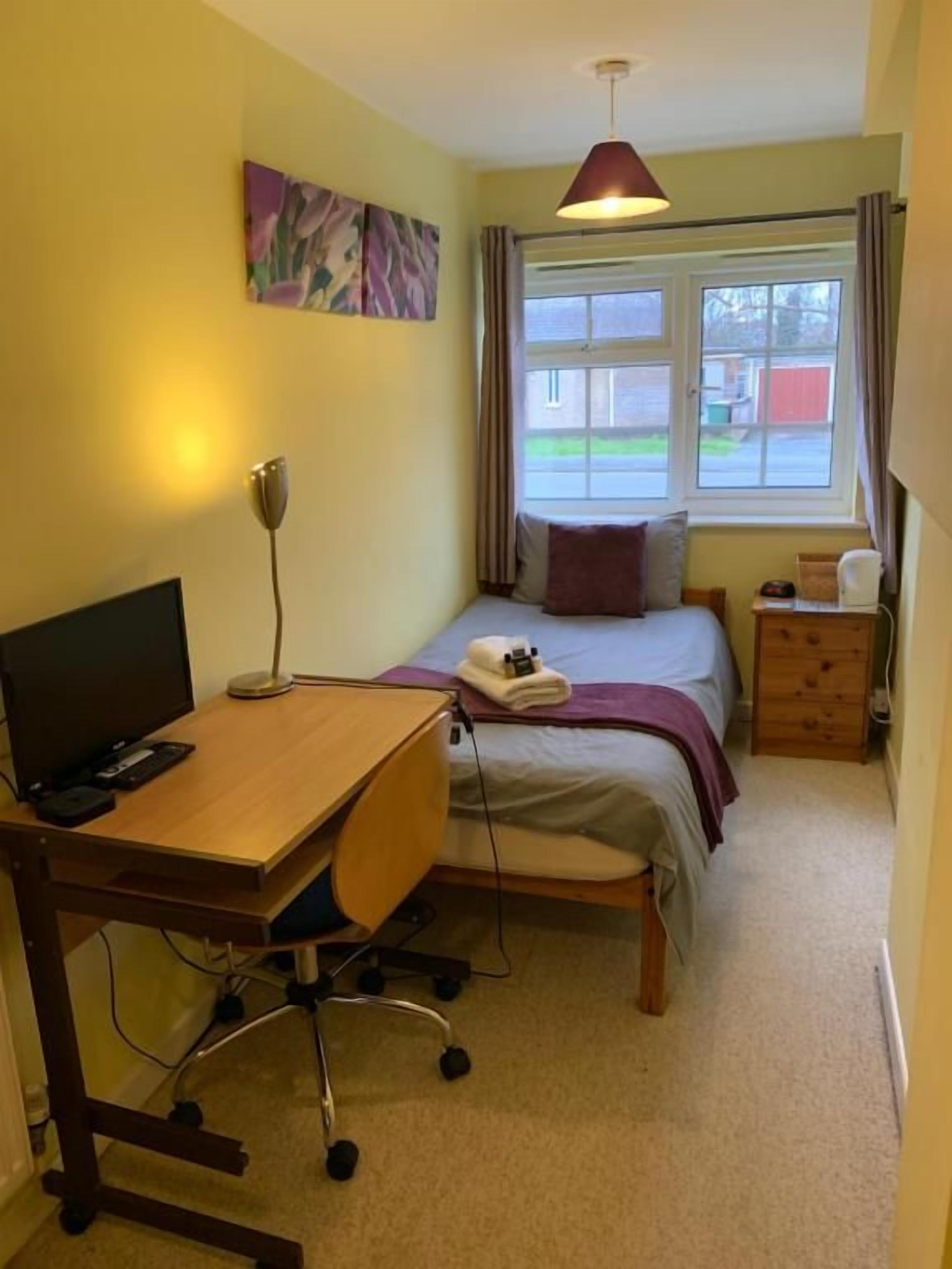 A bedroom with a bed, a desk, a chair, a lamp, and a window.