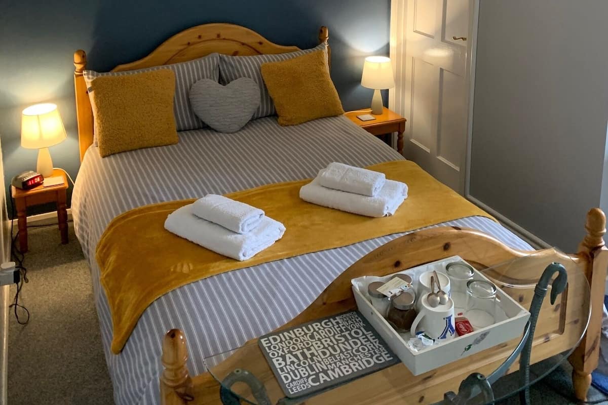 Una cama bien hecha con ropa de cama a rayas y dos almohadas, una mesita de noche con una lámpara, un cuadro colgado en la pared y una bandeja con artículos de aseo.