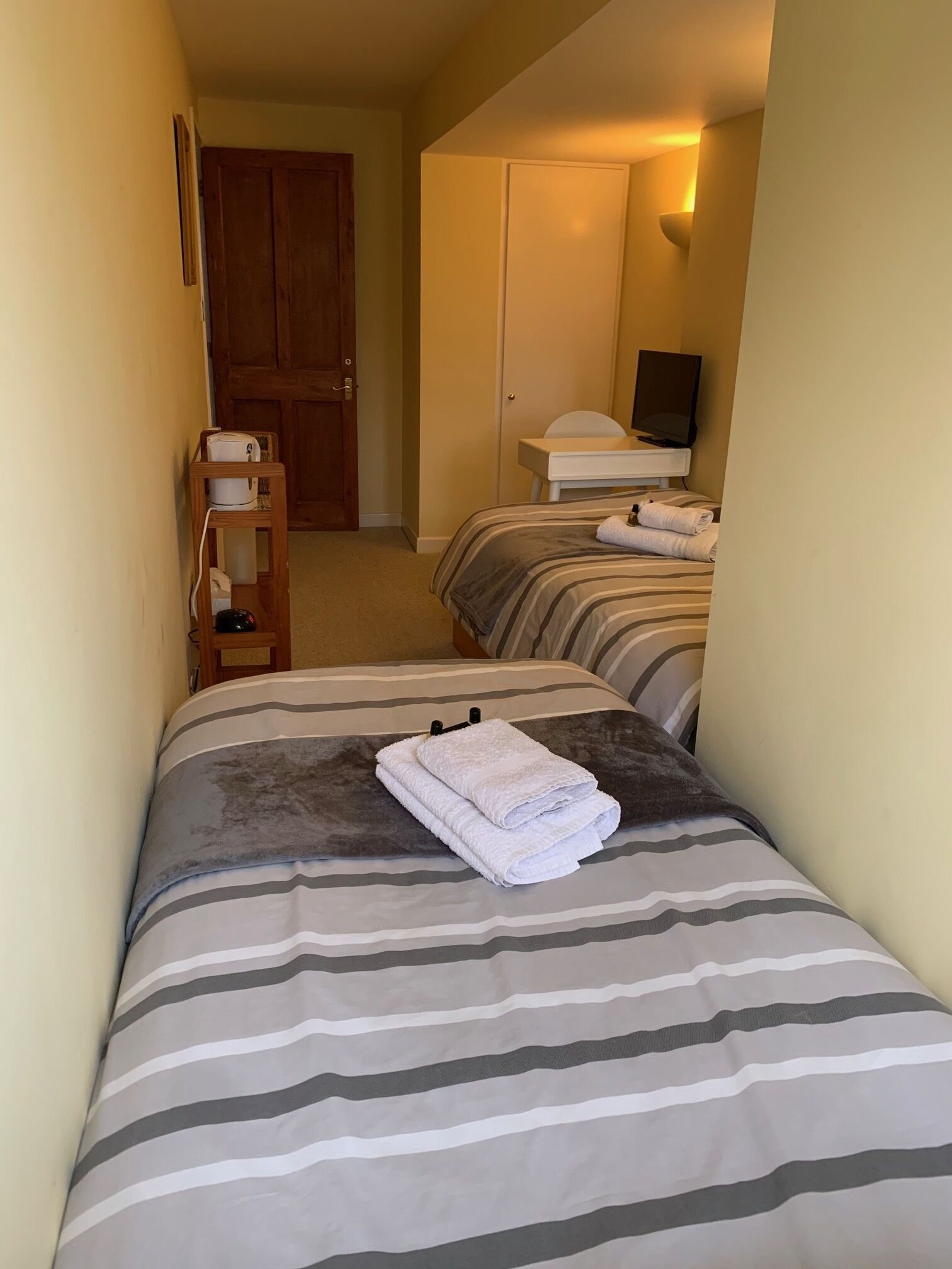 A narrow hotel room with a single bed, a bedside table, and a small desk with a chair.