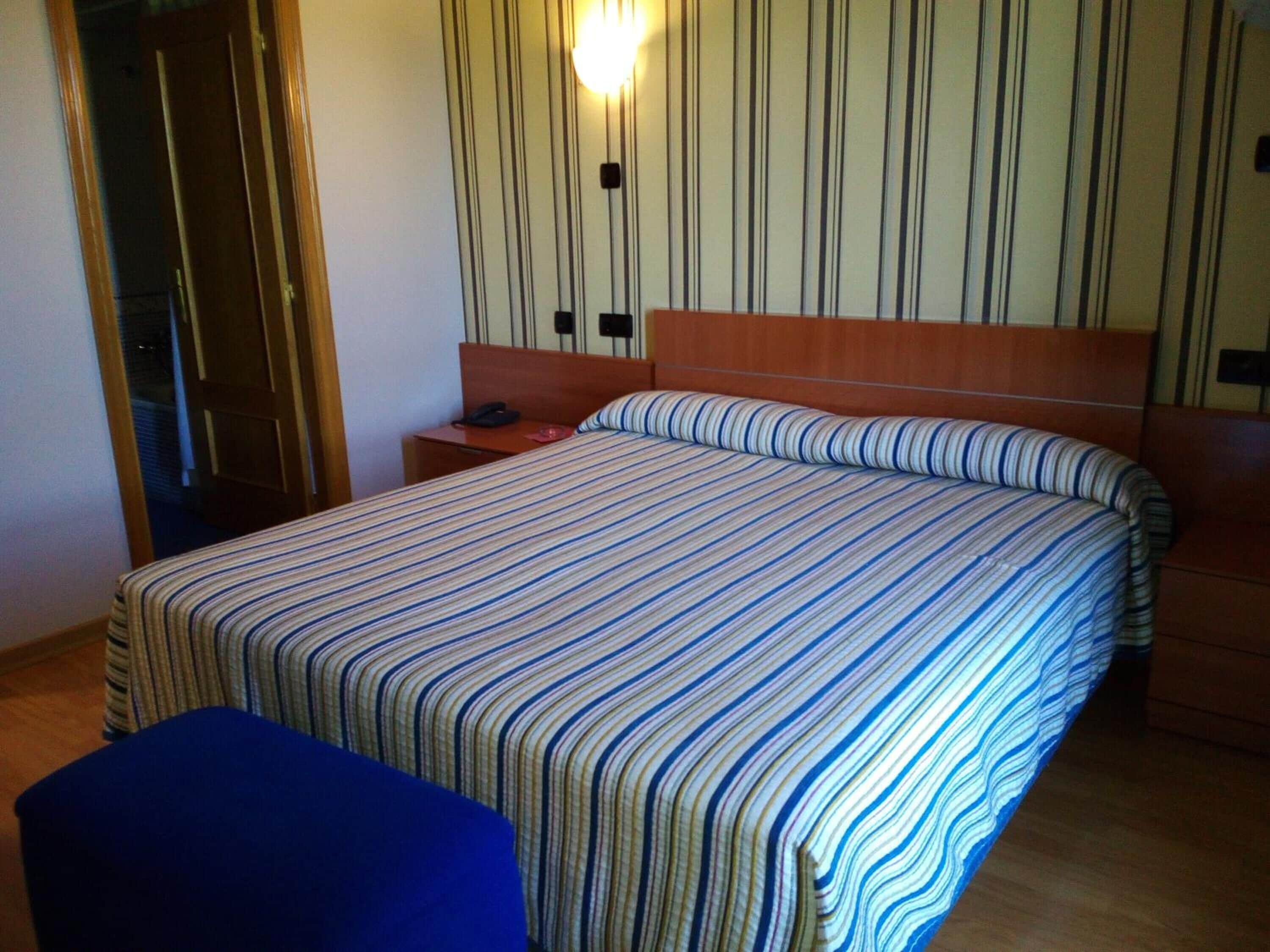 Una habitación de hotel con una cama grande, cabecero de madera, mesita de noche con un teléfono y una lámpara fijada a la pared.