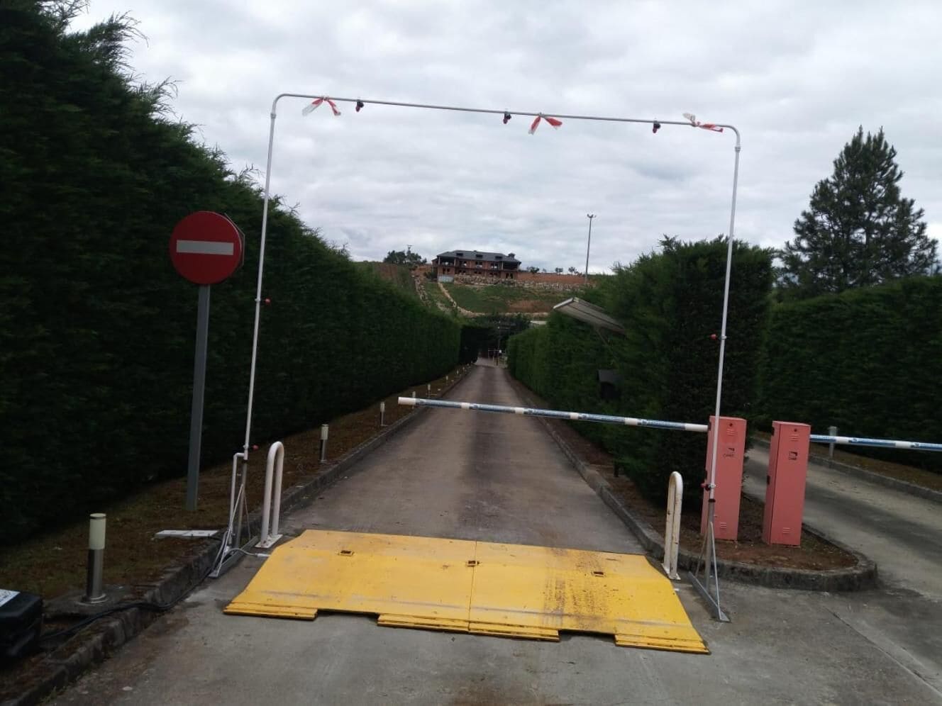 Una carretera que lleva a una entrada vigilada, con un cartel de 'prohibido el paso', una barrera amarilla y una reja roja.