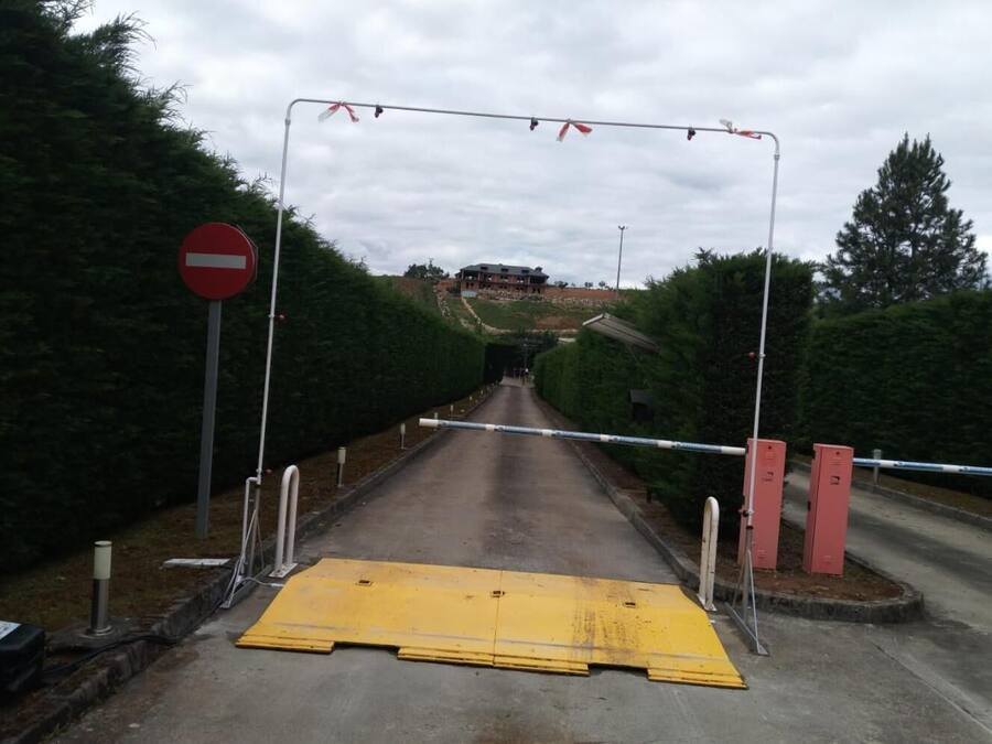 Una carretera que lleva a una entrada vigilada, con un cartel de 'prohibido el paso', una barrera amarilla y una reja roja.