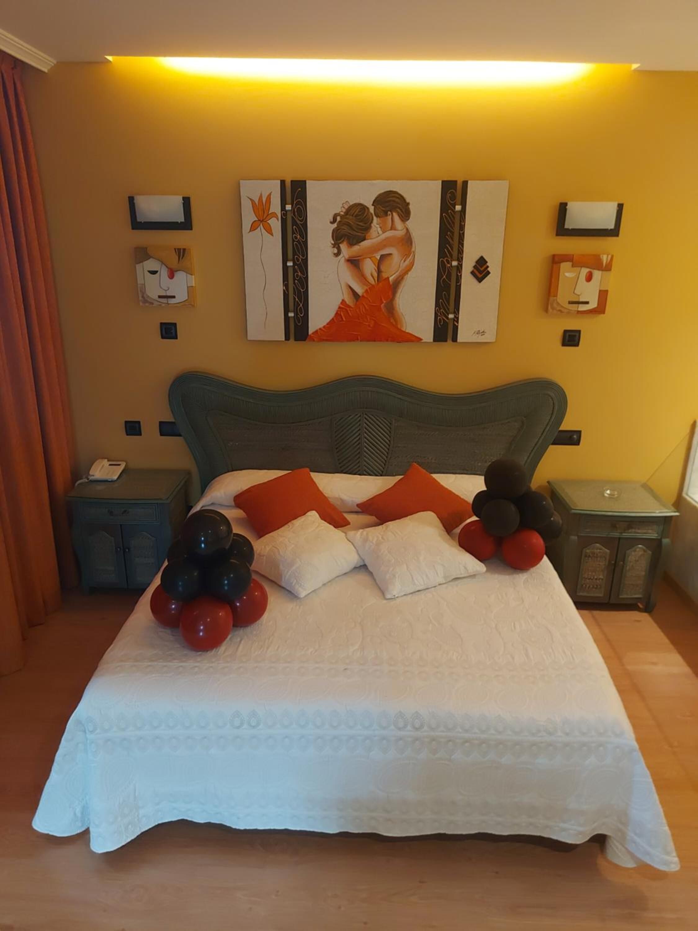 A neatly made bed with a white duvet, red and black balloons, and a painting of a couple on the wall.