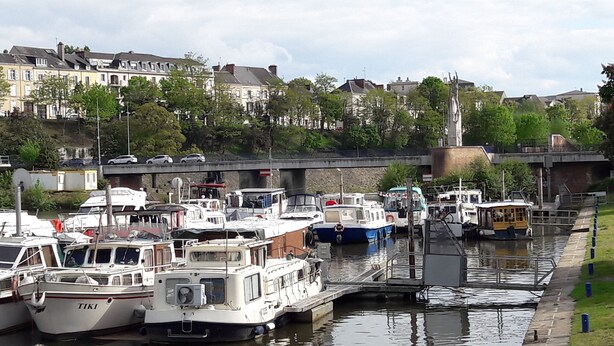 A marina with various boats docked, a bridge in the background, and buildings along the riverbank. - Les Lamartine (Le Mans)