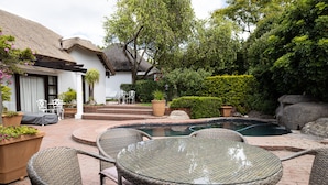 A pool area with wicker chairs and a table, surrounded by a garden with a small pool and a thatched-roof building. - Cape Village Lodge (Cape Town)
