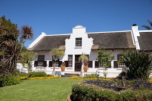 A white colonial-style building with a thatched roof, surrounded by well-maintained gardens and palm trees. - Cape Village Lodge (Cape Town)