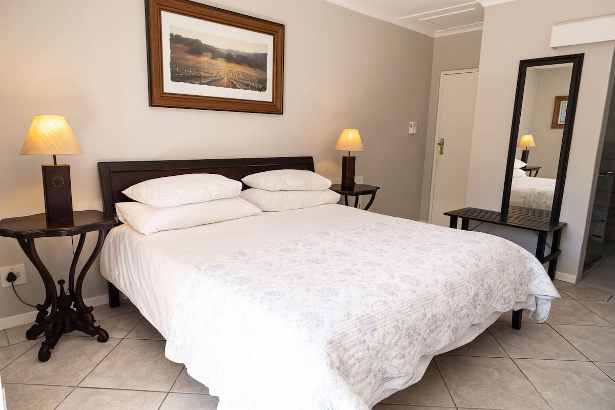A neatly made bed with white linens, two bedside lamps, a framed landscape painting above the bed, and a mirror on the wall.