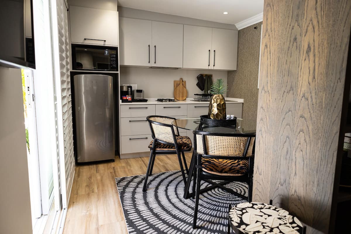 A compact kitchen with white cabinets, a stainless steel refrigerator, a microwave, and a coffee maker. There are two wicker chairs with patterned cushions around a small table.