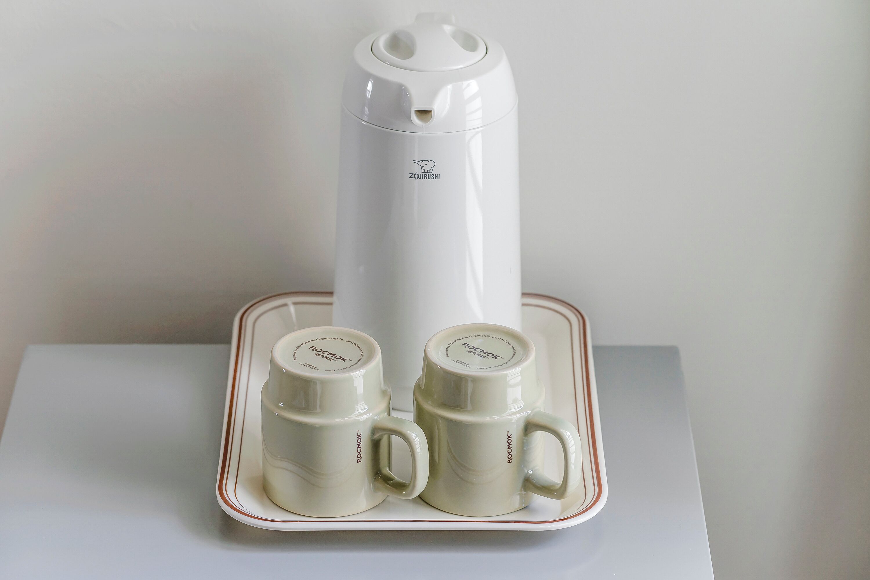 A white electric kettle with a logo, two white mugs with a handle, and a tray with a red border.
