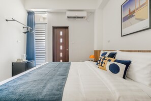 Une chambre d’hôtel comprenant un lit, une table de chevet, un climatiseur fixé au mur, une porte et une fenêtre avec des rideaux.