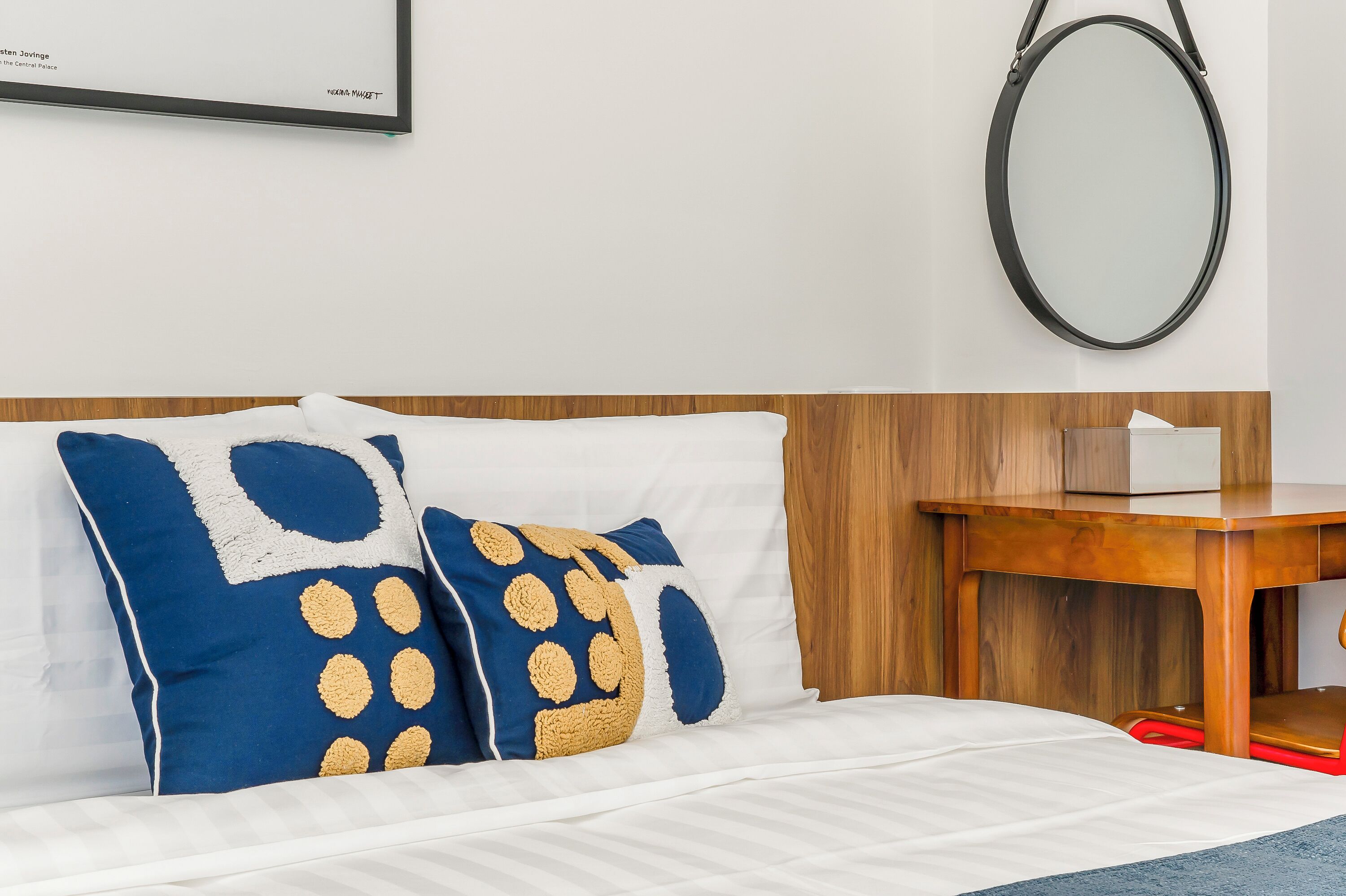 A neatly made bed with white linens, two decorative pillows with blue and beige patterns, a wooden nightstand with a tissue box, and a round mirror on the wall.