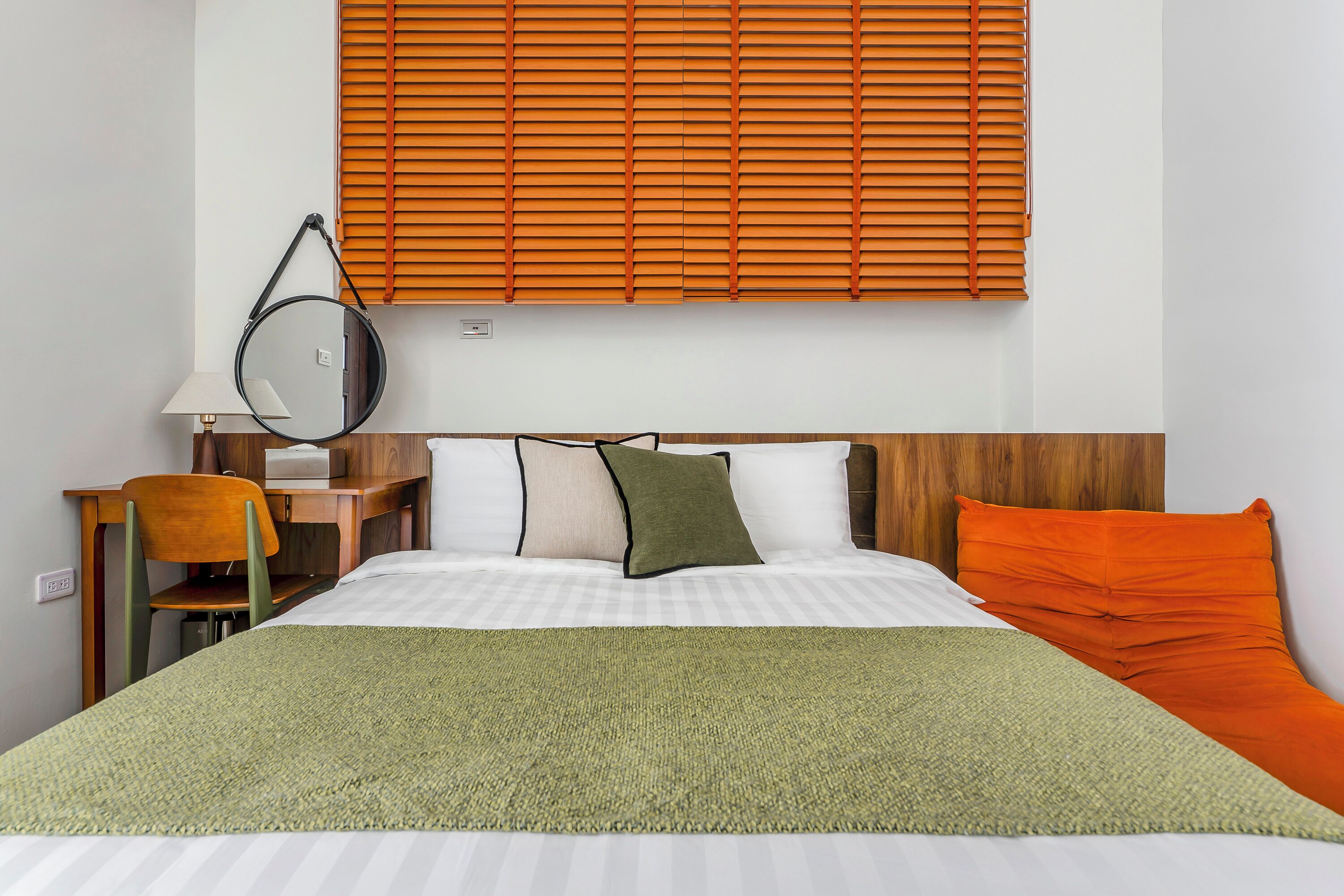 A hotel room with a bed, a desk with a chair, a lamp, and a window with blinds.