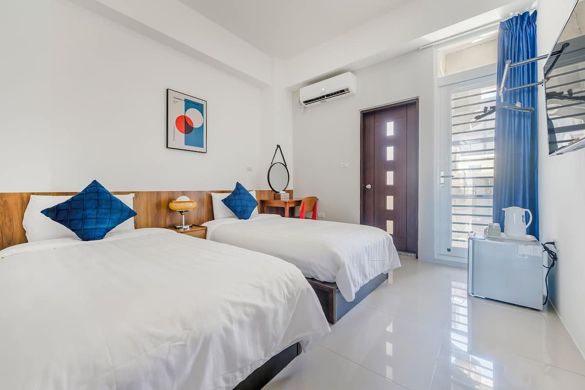 Une chambre d’hôtel avec deux lits, une tête de lit en bois, une petite table avec une lampe, un climatiseur fixé au mur et une porte donnant sur un balcon avec vue.