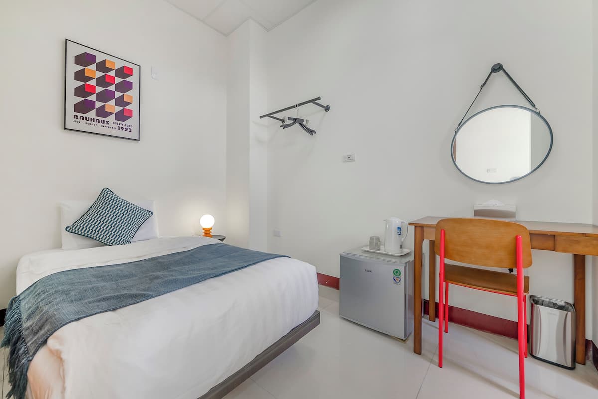 Une chambre d’hôtel équipée d’un lit, d’un bureau, d’une chaise, d’un petit radiateur, d’une poubelle et d’un miroir fixé au mur.