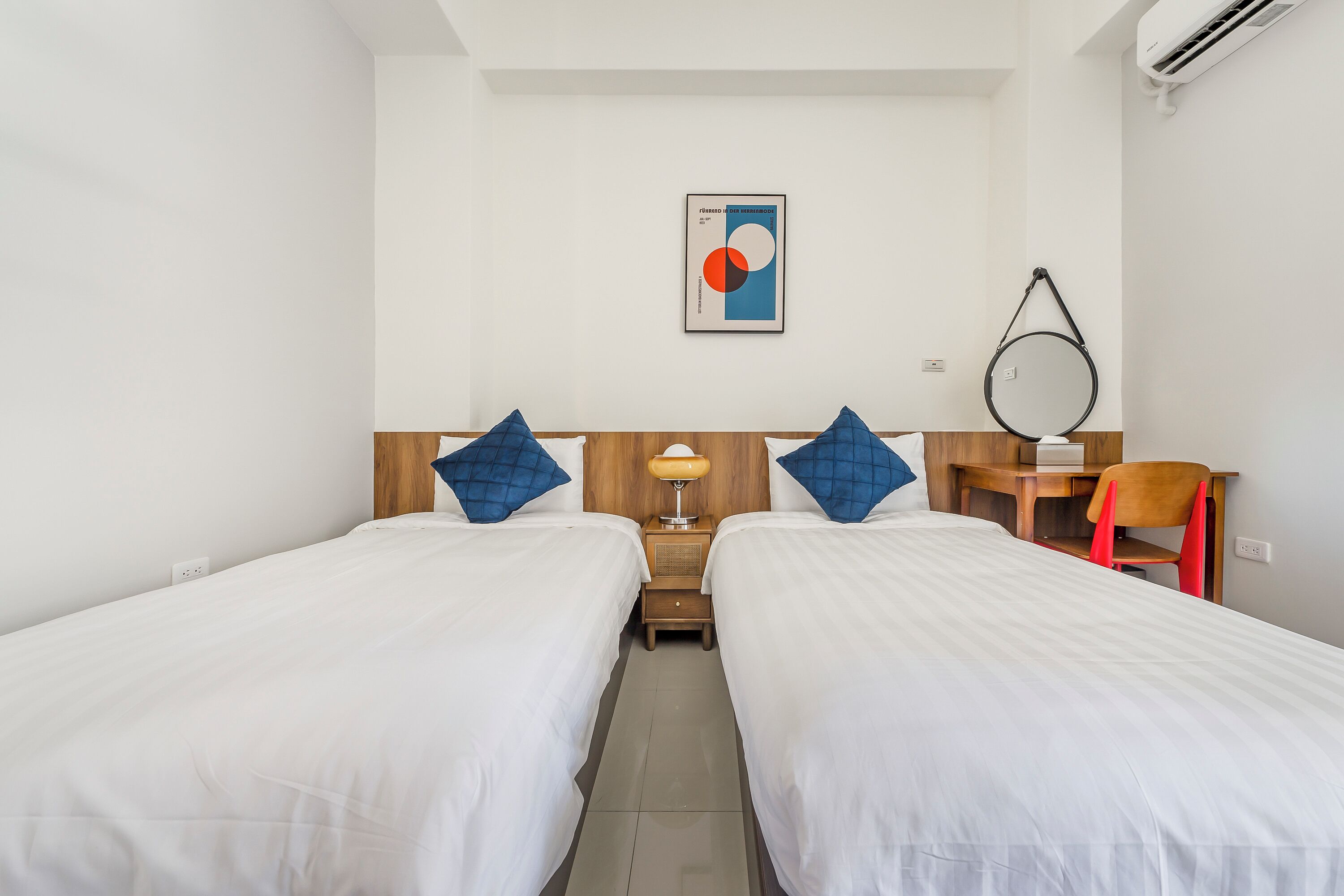 Quarto de hotel com duas camas de solteiro, cabeceira de madeira, pequena escrivaninha, cadeira, luminária e espelho fixado na parede.