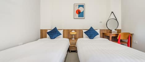 Quarto de hotel com duas camas de solteiro, cabeceira de madeira, pequena escrivaninha, cadeira, luminária e espelho fixado na parede.