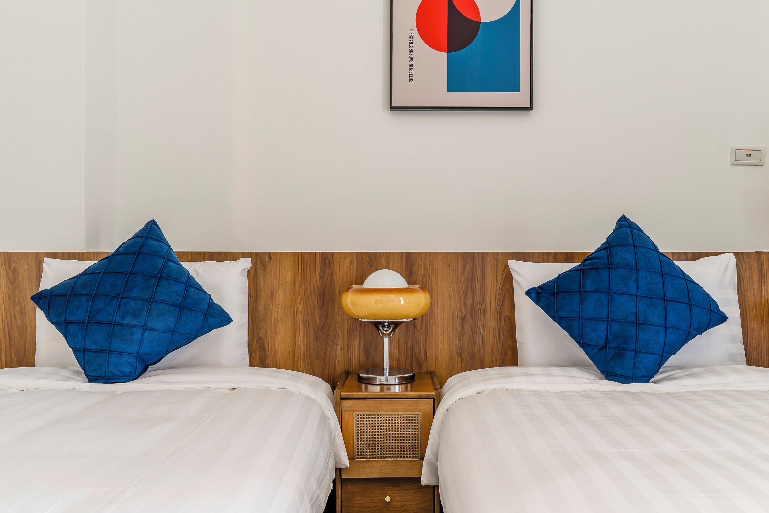 A hotel room with two beds, each with a blue pillow and white bedding. A wooden headboard separates the beds, and a small table with a lamp is placed between them.