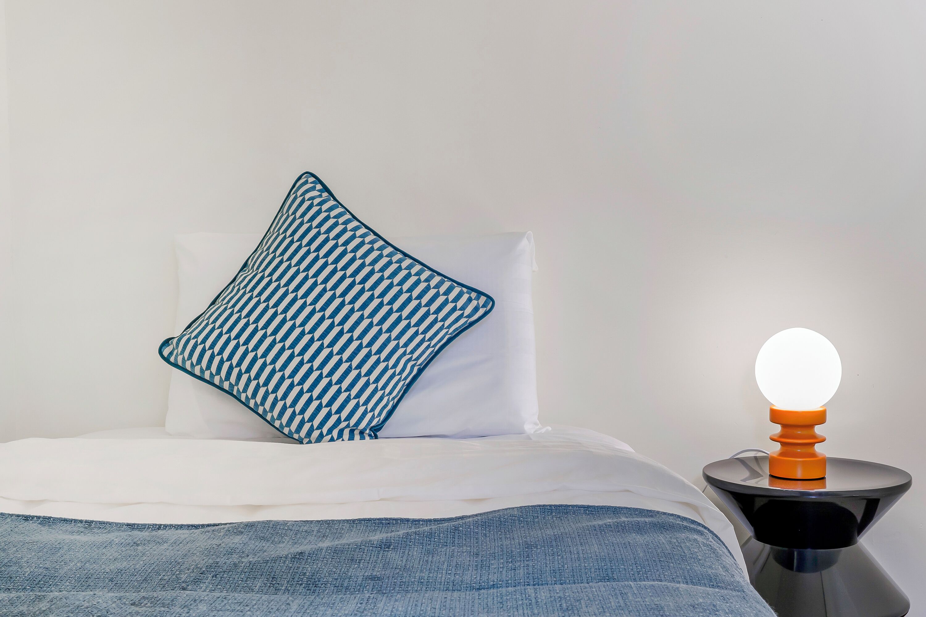 A bed with white bedding and a blue patterned pillow, a black nightstand with a round orange lamp, and a plain white wall.