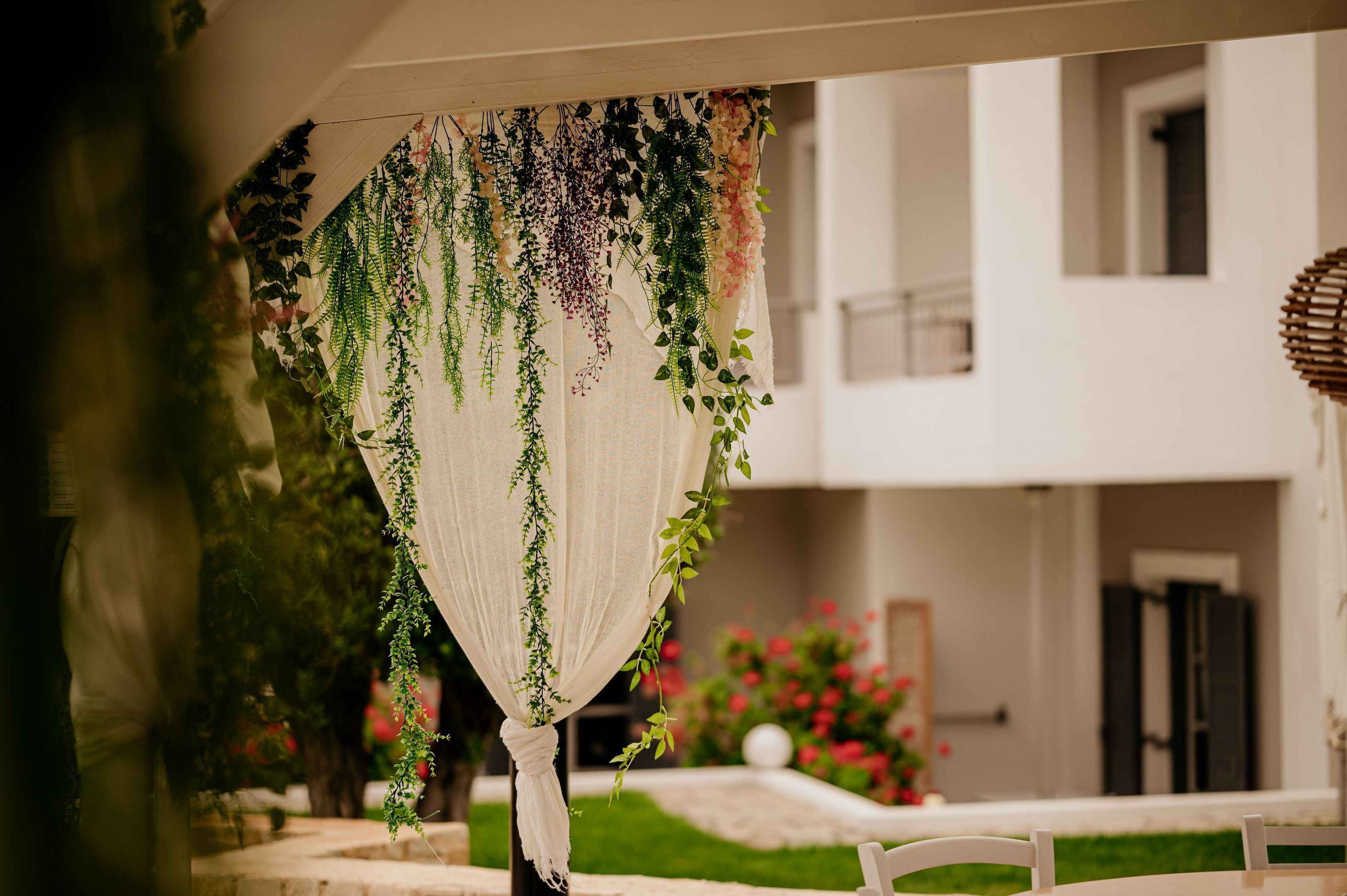 An outdoor area with a white chair, a hanging plant arrangement, and a building in the background.
