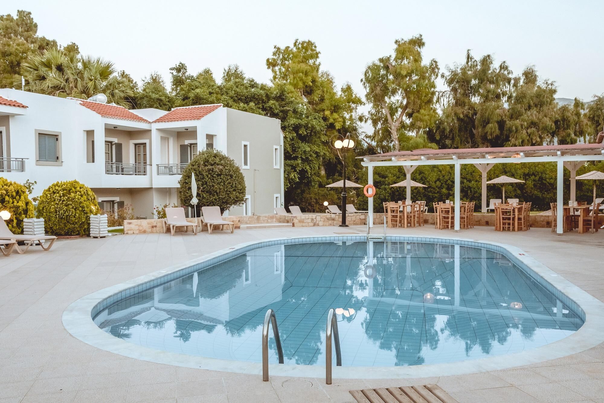 A swimming pool with lounge chairs and umbrellas, surrounded by a building with a red-tiled roof and a covered outdoor seating area.
