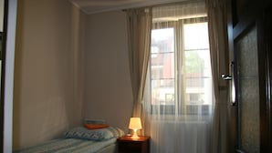 Een kleine, eenvoudige slaapkamer met een enkel bed, een nachtkastje met een lamp en een raam met dunne gordijnen.