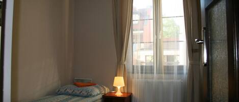 Un dormitorio pequeño y sencillo con una cama individual, una mesita de noche con lámpara y una ventana con cortinas traslúcidas.