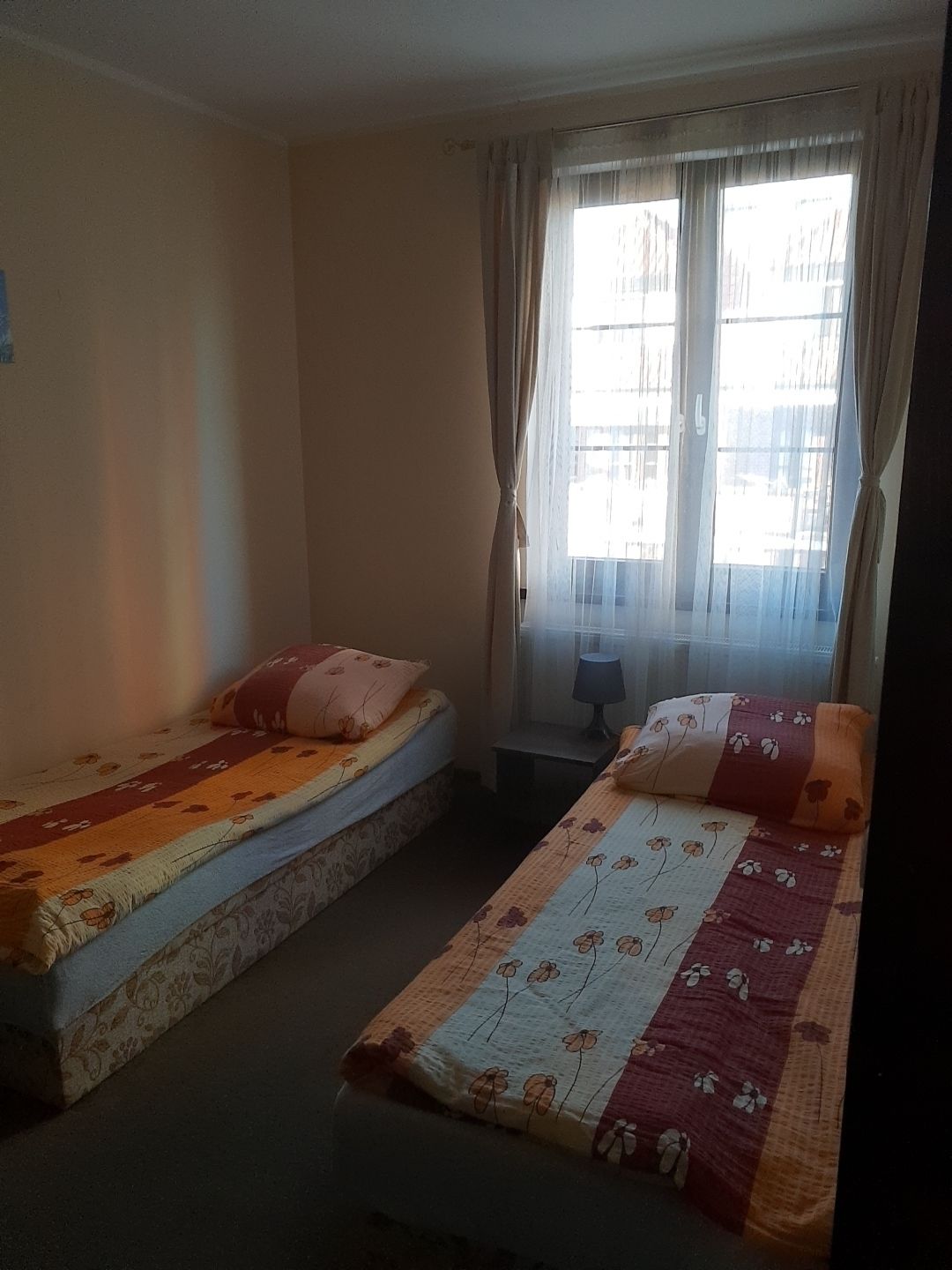 Une chambre avec deux lits simples, chacun agrémenté de draps à motifs floraux. Une fenêtre, ornée de rideaux légers, offre une vue sur les bâtiments environnants.
