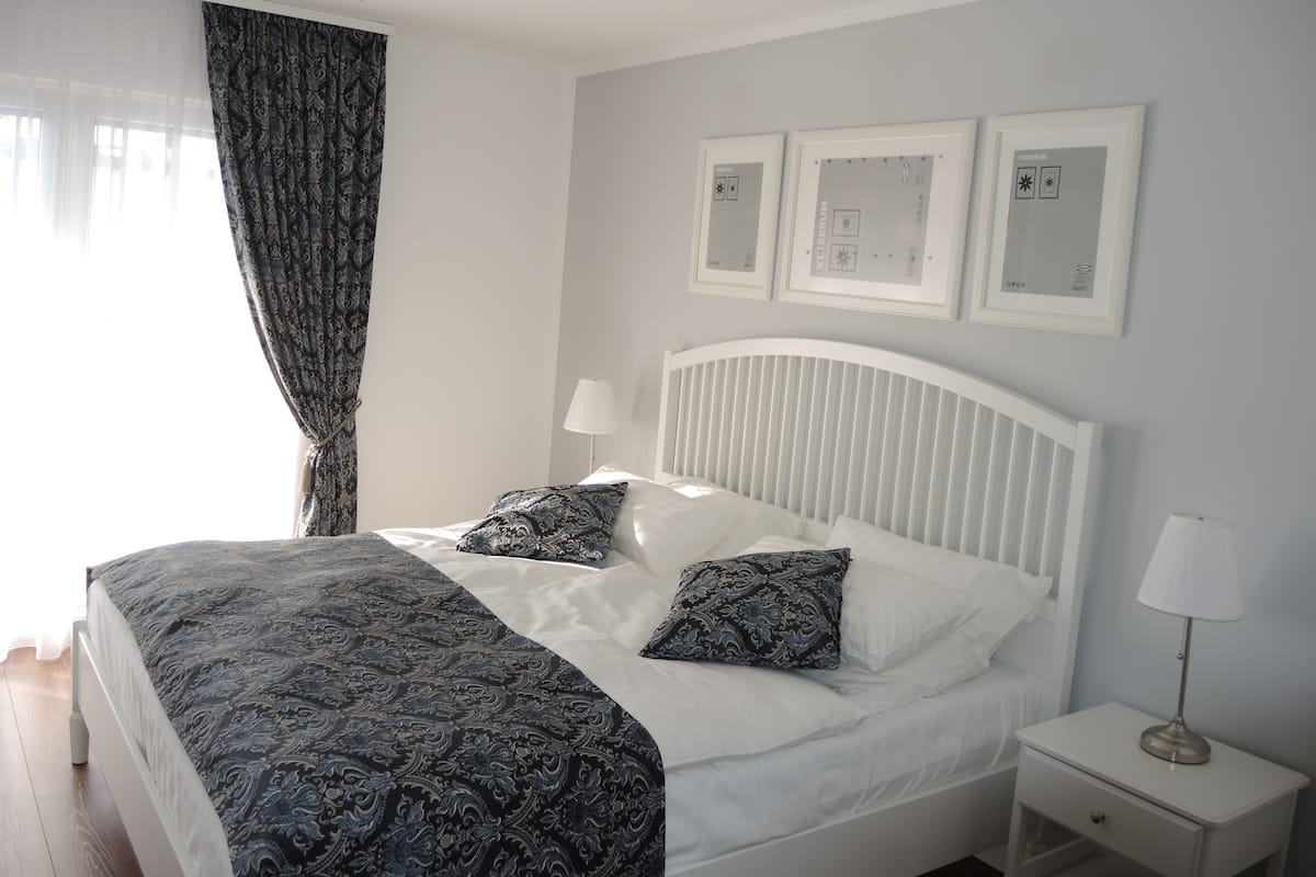 A neatly made bed with a patterned bedspread and pillows, a bedside table with a lamp, and a window with sheer curtains.