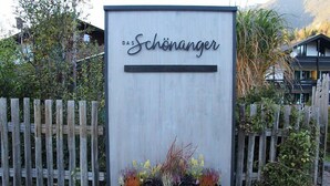 Un letrero con el texto 'DAS Schönanger' en una fuente moderna, sobre un fondo de madera y con una jardinera en la parte inferior.