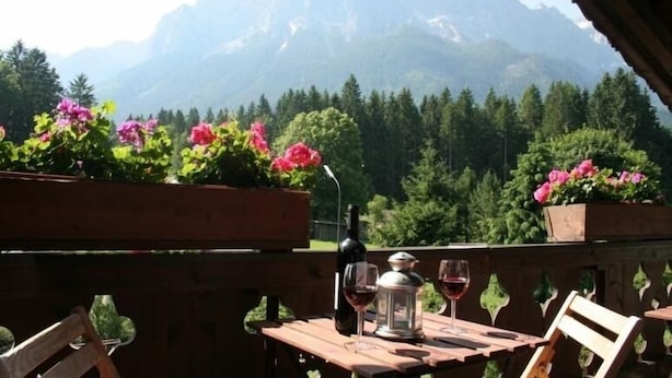 Una mesa de madera con una botella y copas, rodeada de macetas con flores y una vista a la montaña.