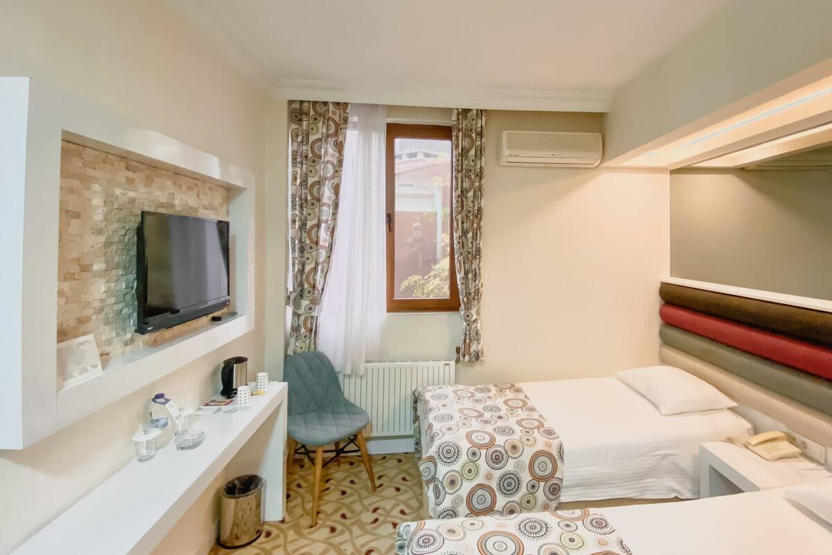 A hotel room with a TV mounted on the wall, a desk with a chair, a small window with curtains, a radiator, and two beds with patterned bedding.