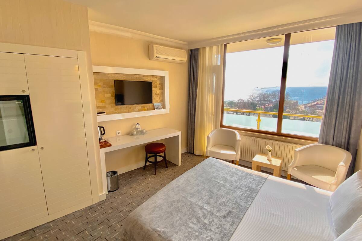 A hotel room with a bed, a desk, a chair, a TV, and a view of the sea.