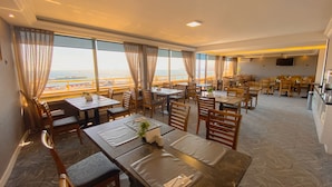 Un restaurante con varias mesas y sillas, ventanas amplias con cortinas y vista al mar.