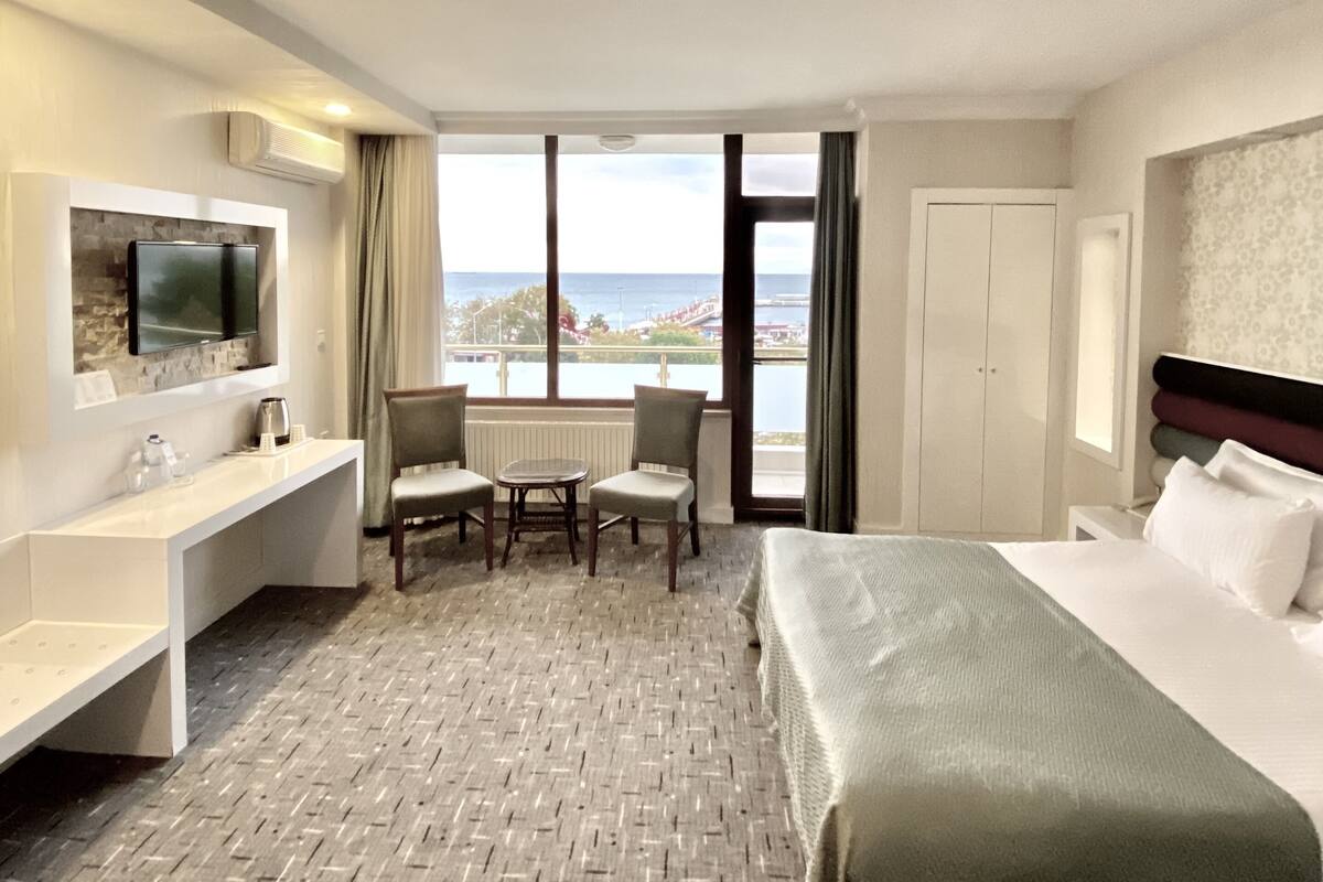 A hotel room with a bed, a desk, a chair, a TV, and a view of the sea.