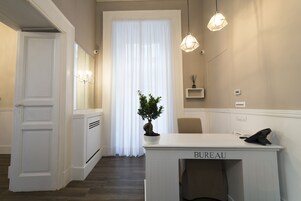 Een hotelkamer met een witte kaptafel, een plant in pot, een stoel en een groot raam met transparante gordijnen.