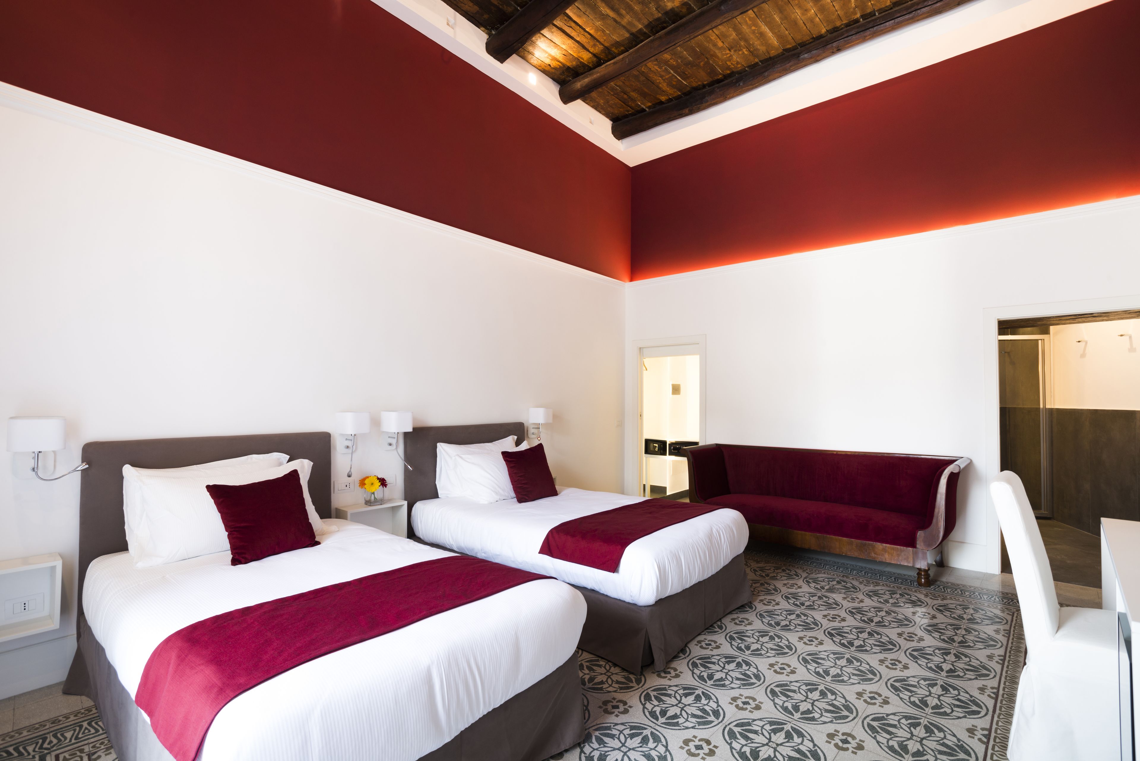 A hotel room with two beds, a red accent wall, patterned floor tiles, and a wooden ceiling beam.