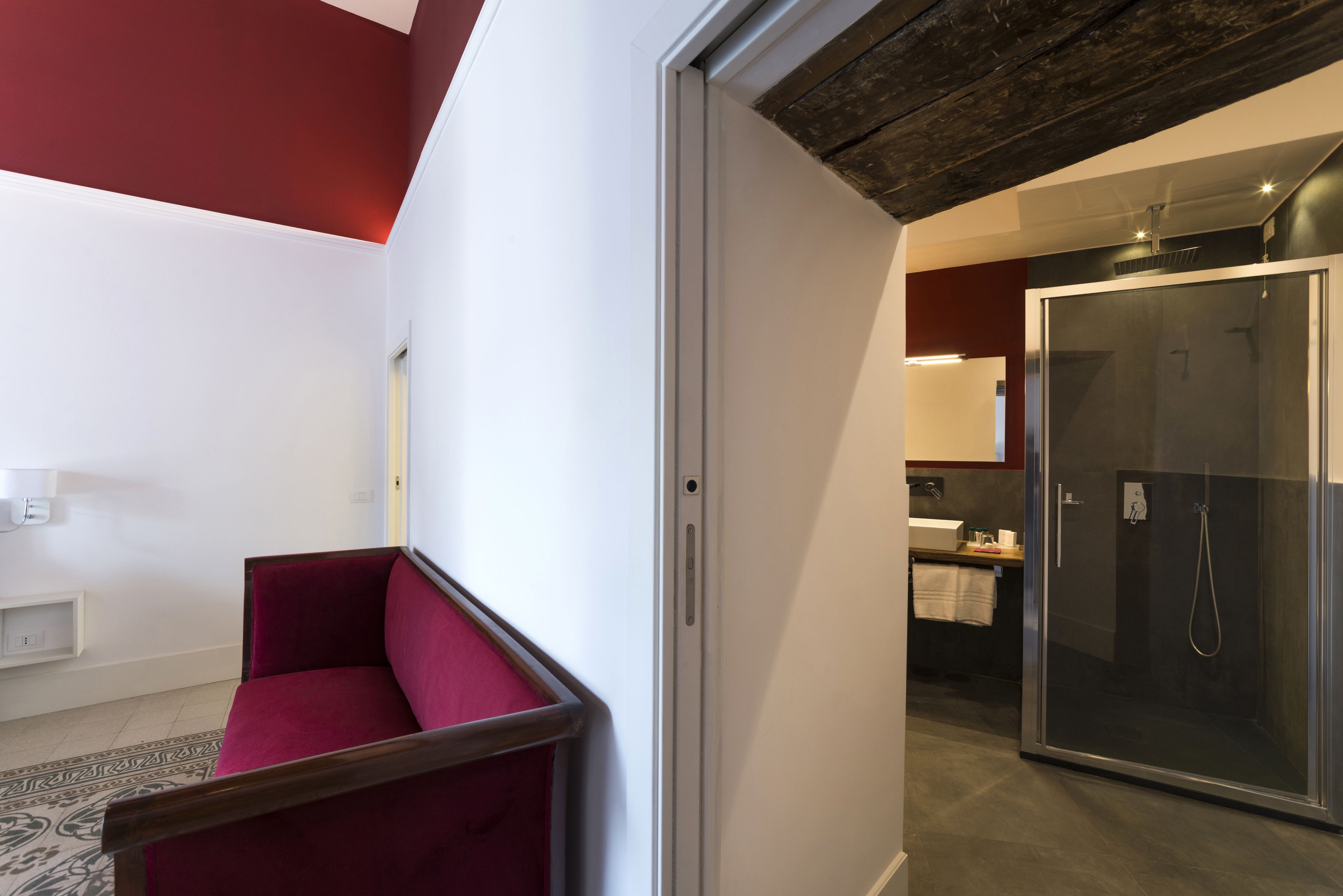 A modern hotel room with a red sofa, a glass shower enclosure, and a kitchen area visible through an open door.